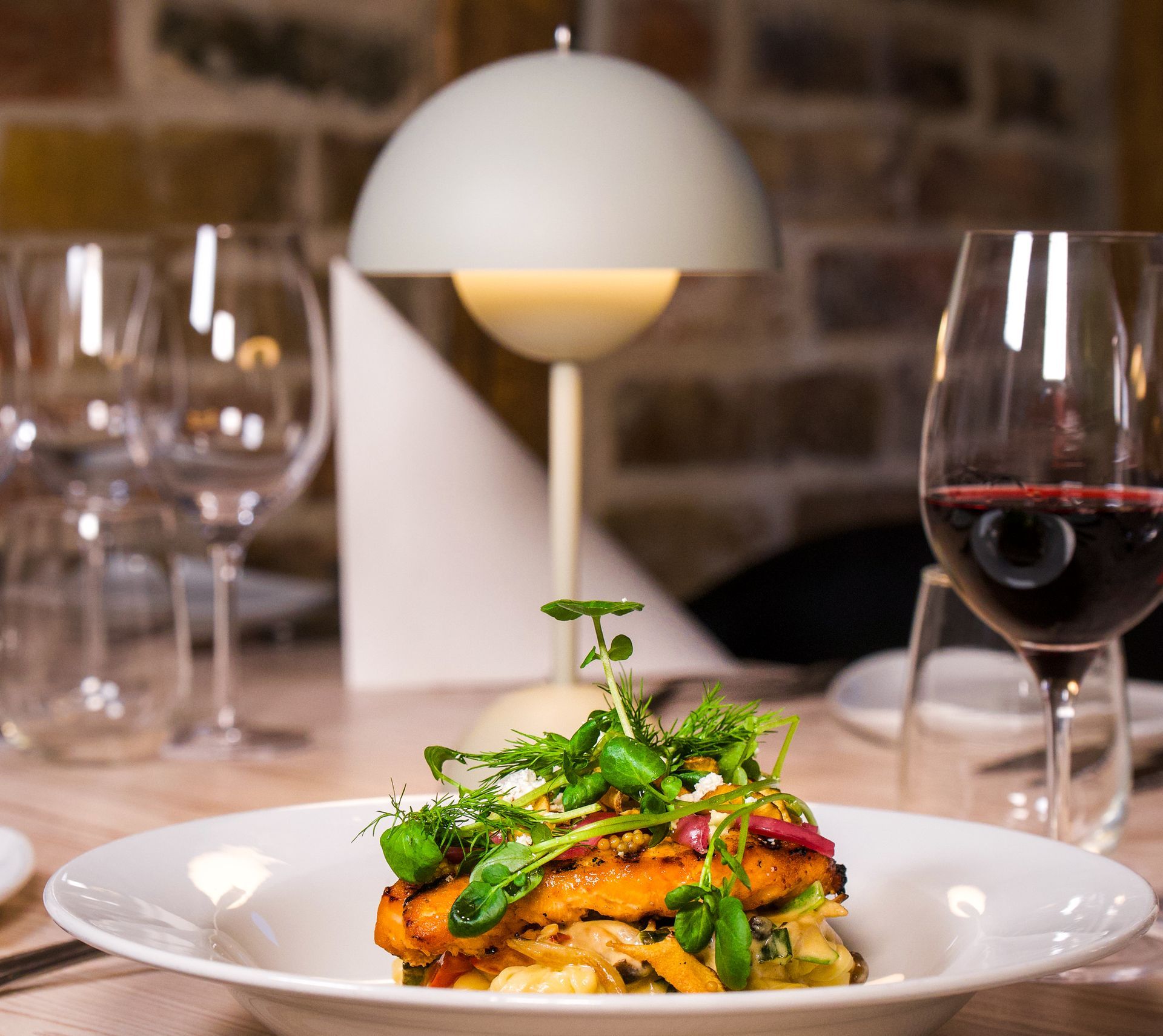 Tallerken med mad på et bord med et vinglas og en lampe; restaurantindstilling.