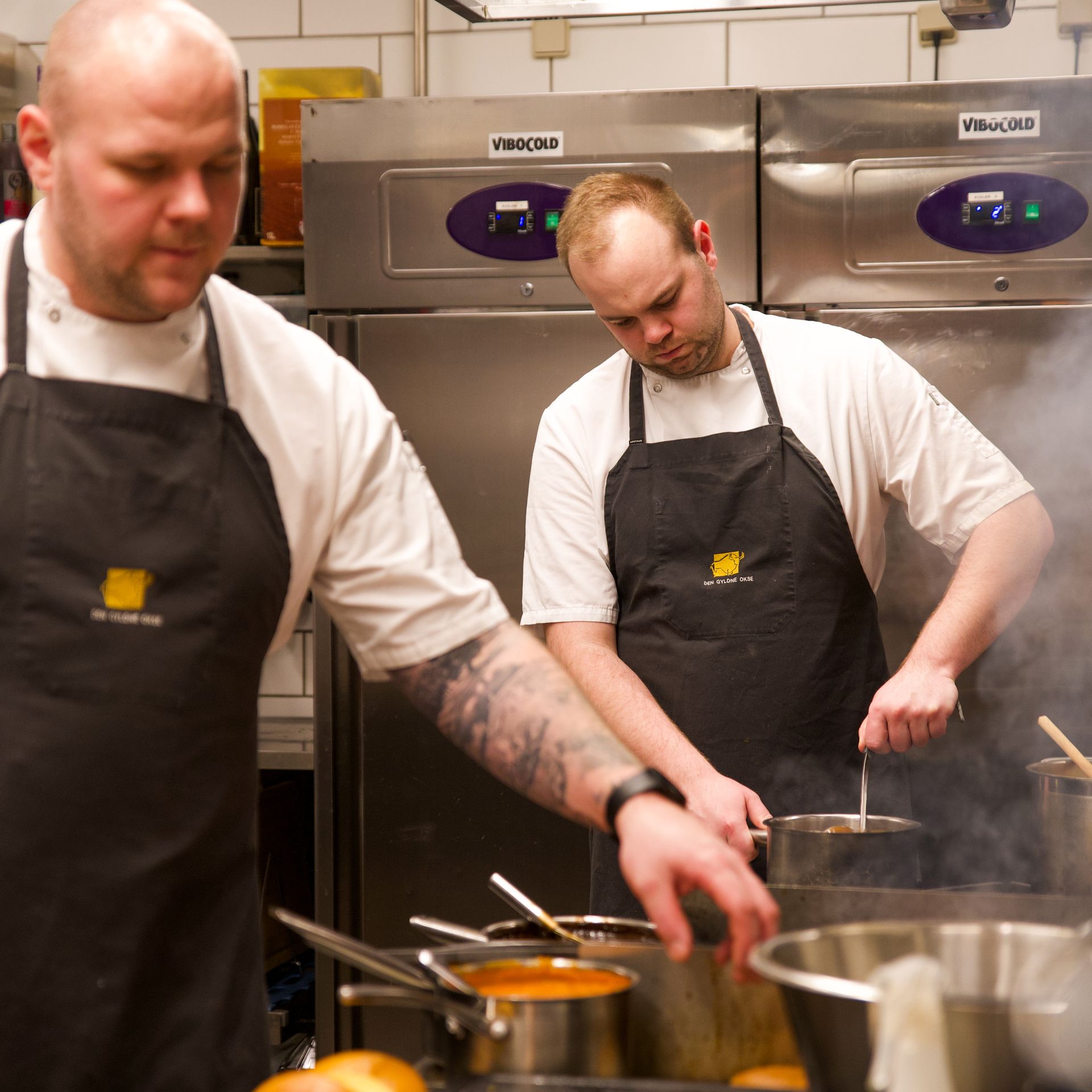 To kokke i hvide skjorter og sorte forklæder laver mad i et restaurantkøkken.