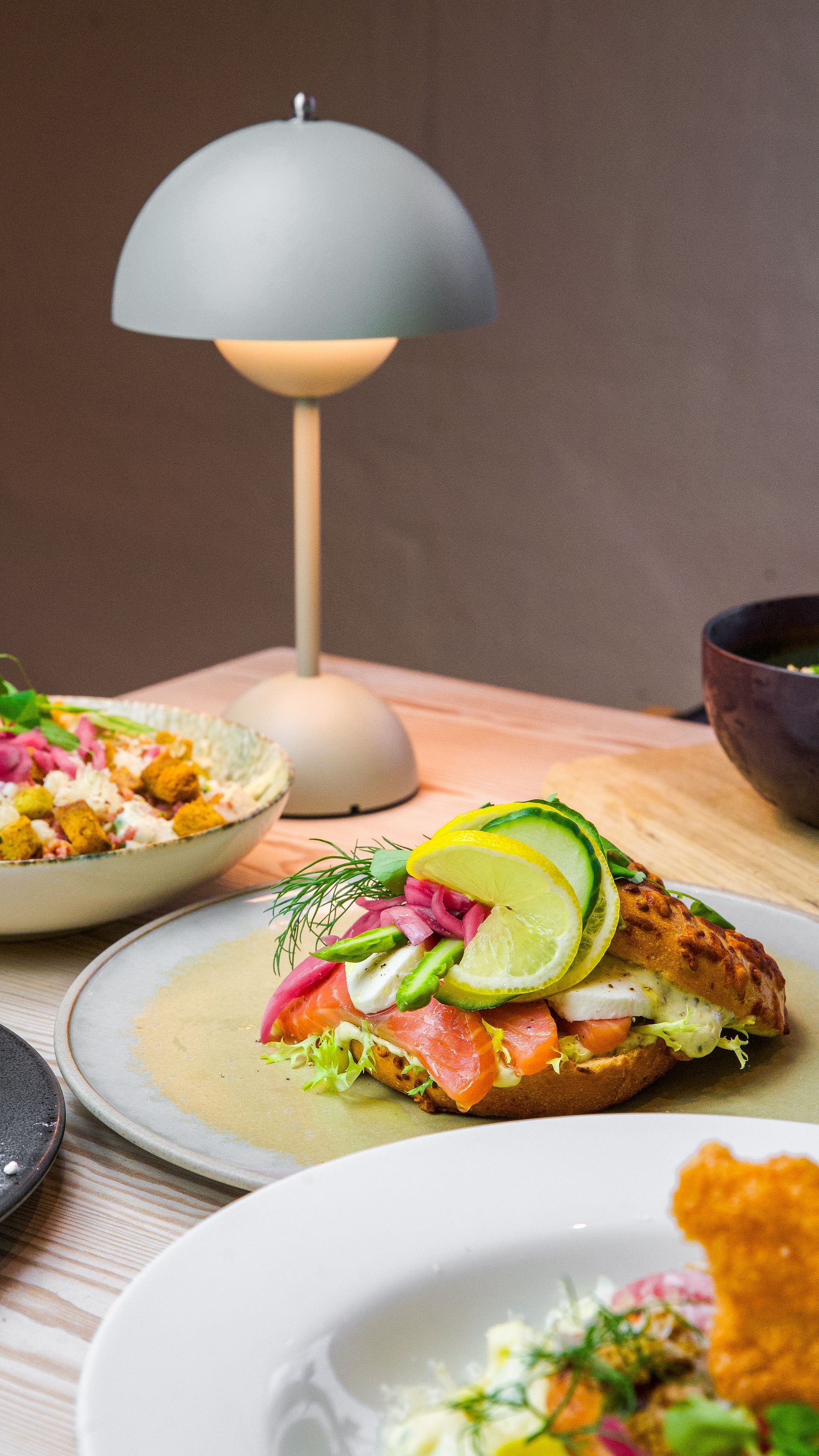 Et bord med mad, en lampe og en laksesandwich med topping.