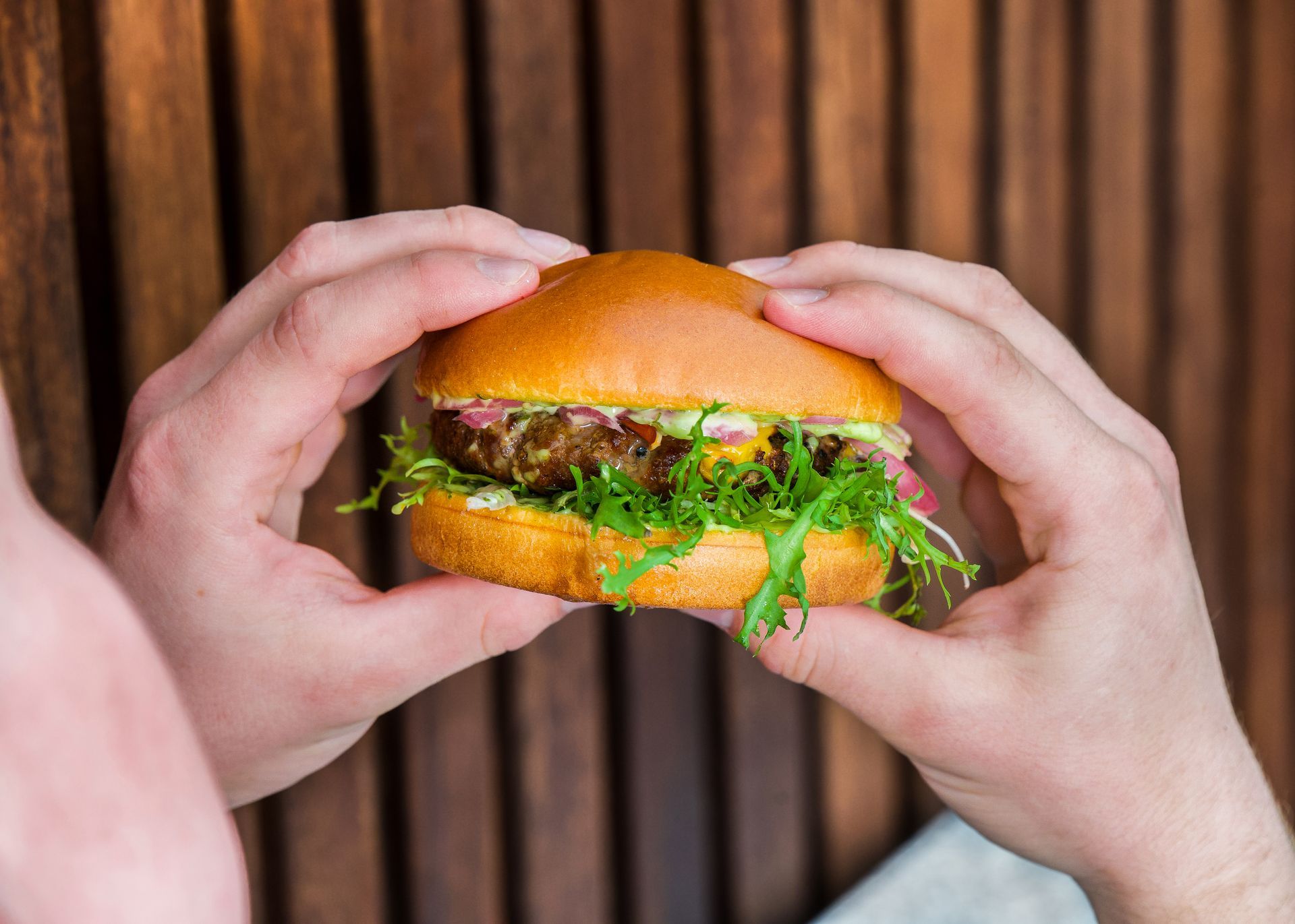 Person, der holder en burger med bolle, salat og bøf mod en træbaggrund.