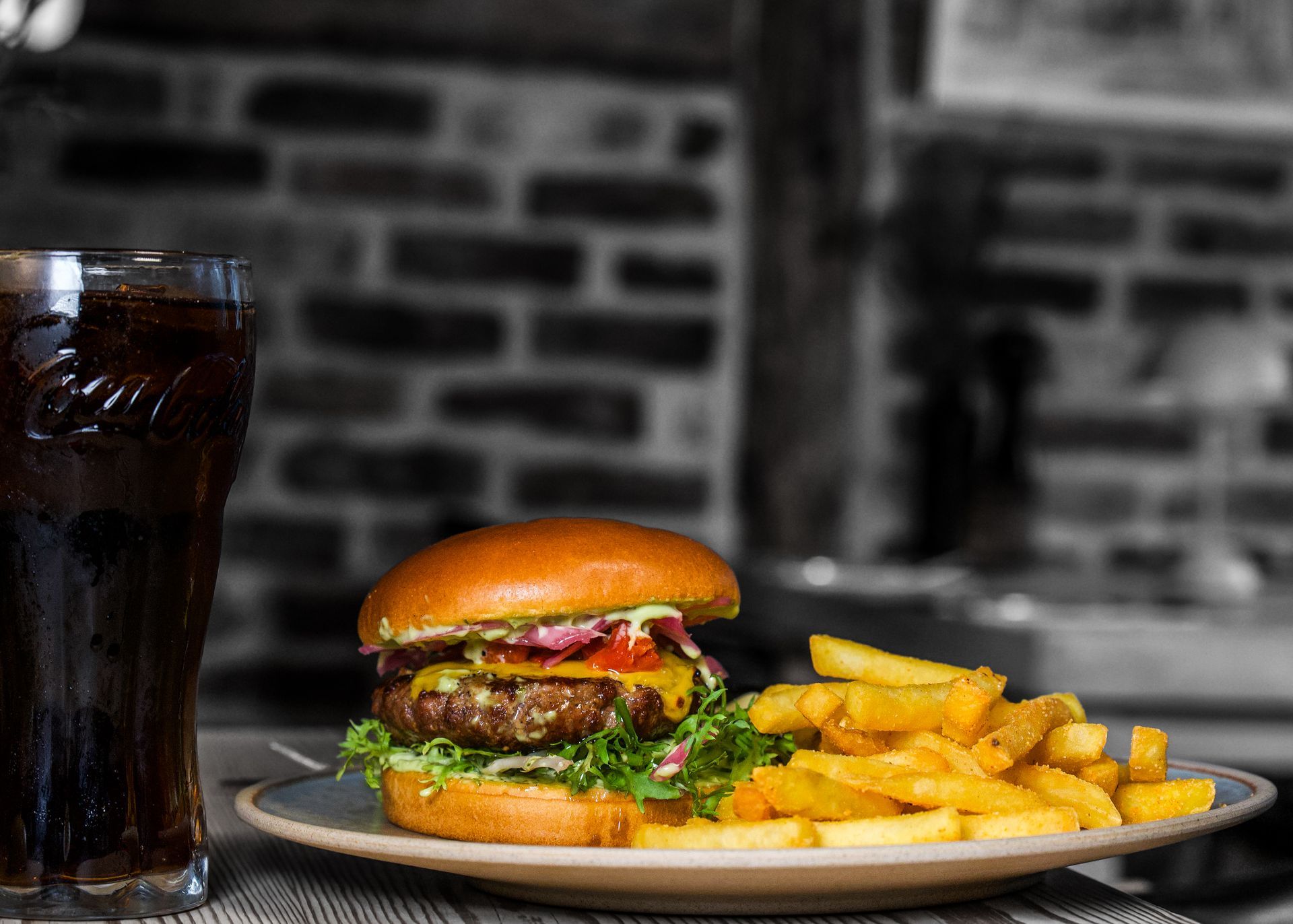 Burger, pommes frites og sodavand på en tallerken, placeret op mod en murstensvæg, oplyst for at skabe en restaurantstemning.