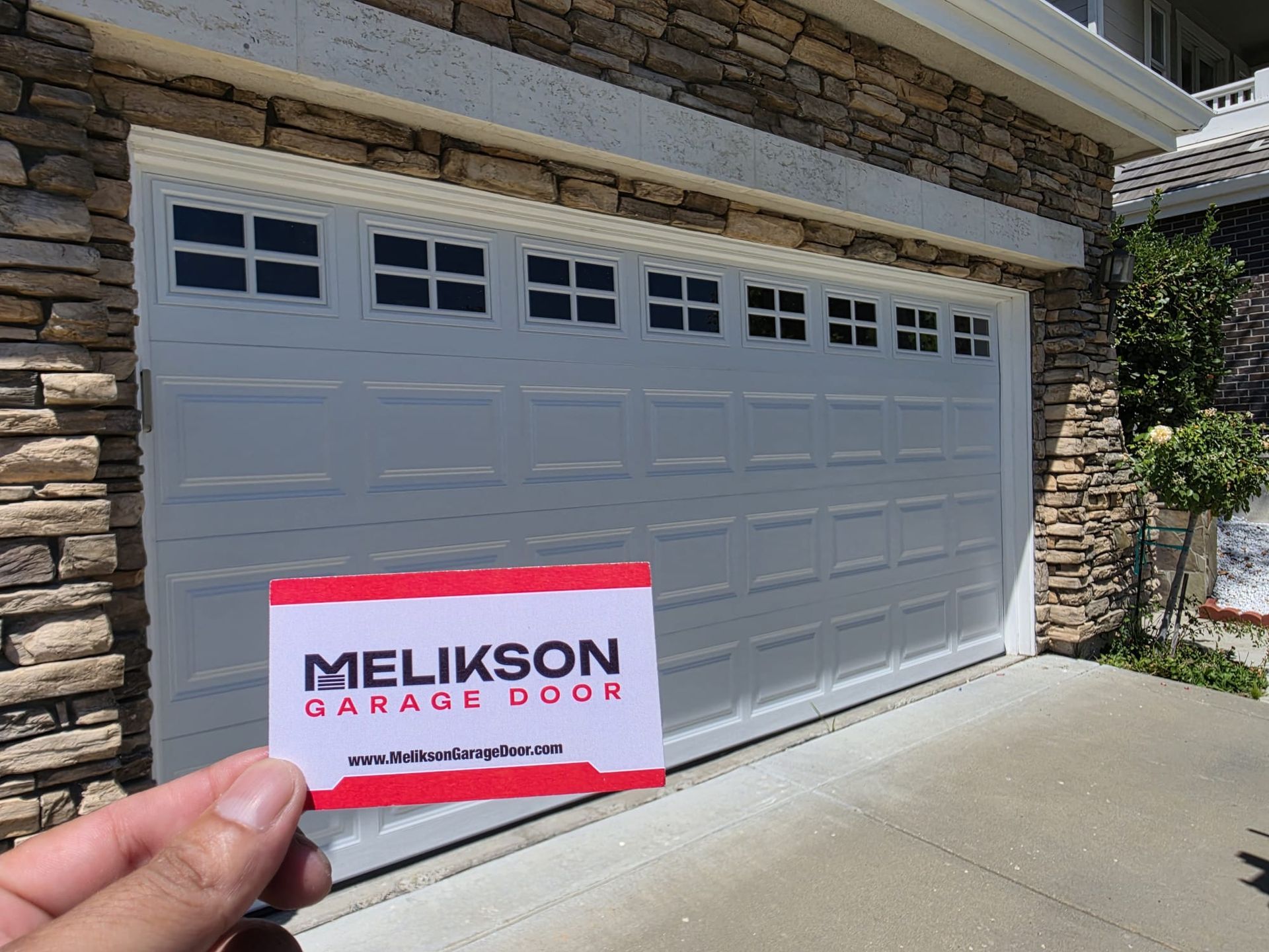 Melikson Garage Door business card held in front of a white raised-panel garage door on a Santa Clara County home