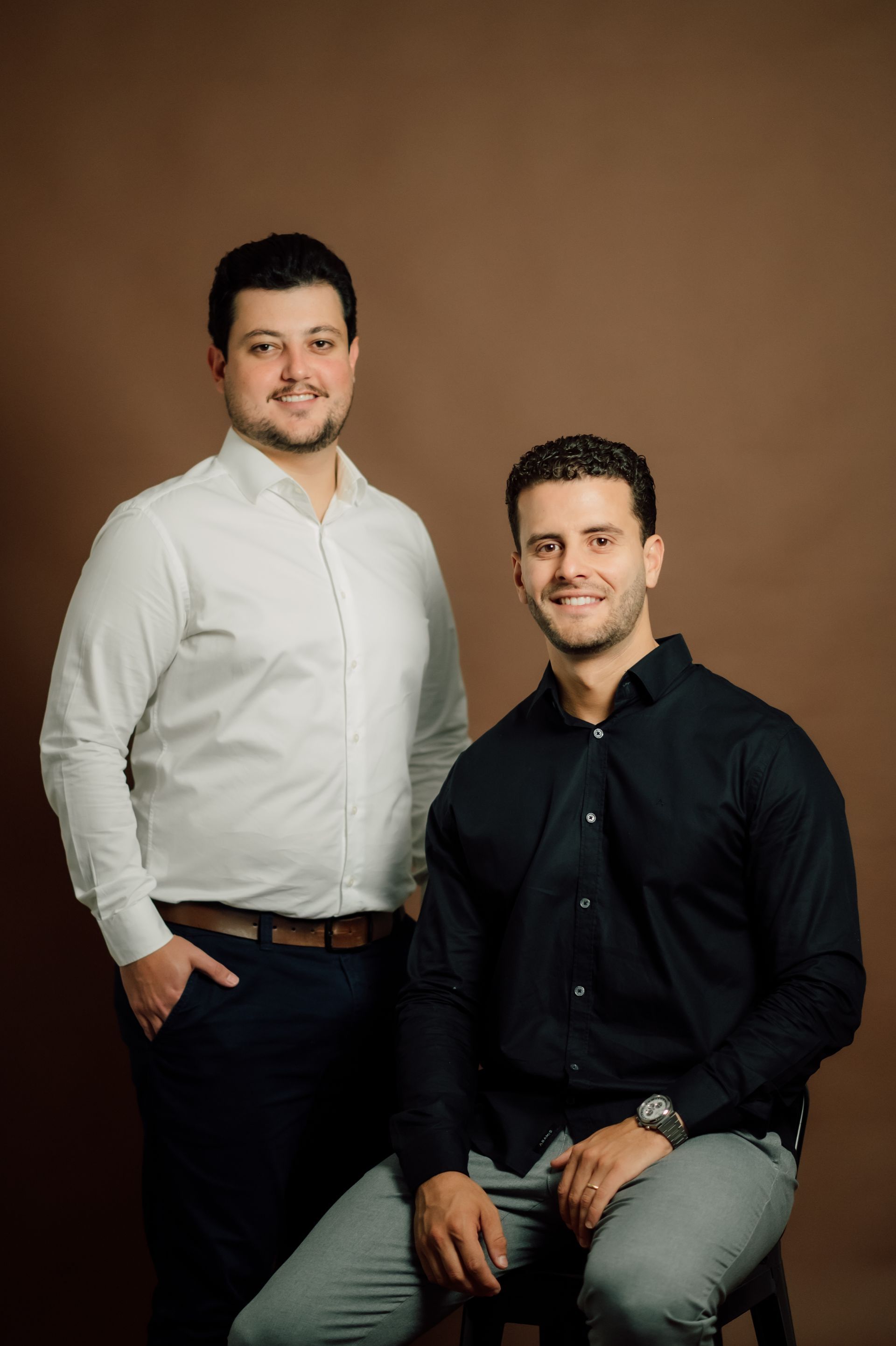 Dois homens posando para retratos de estúdio, um em pé com camisa branca e o outro sentado com camisa preta.
