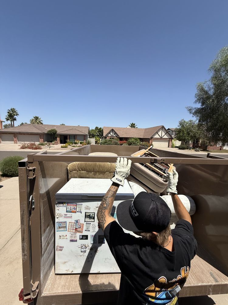 Worker lifting a dumpster lid beside a white utility box in a sunny suburban street