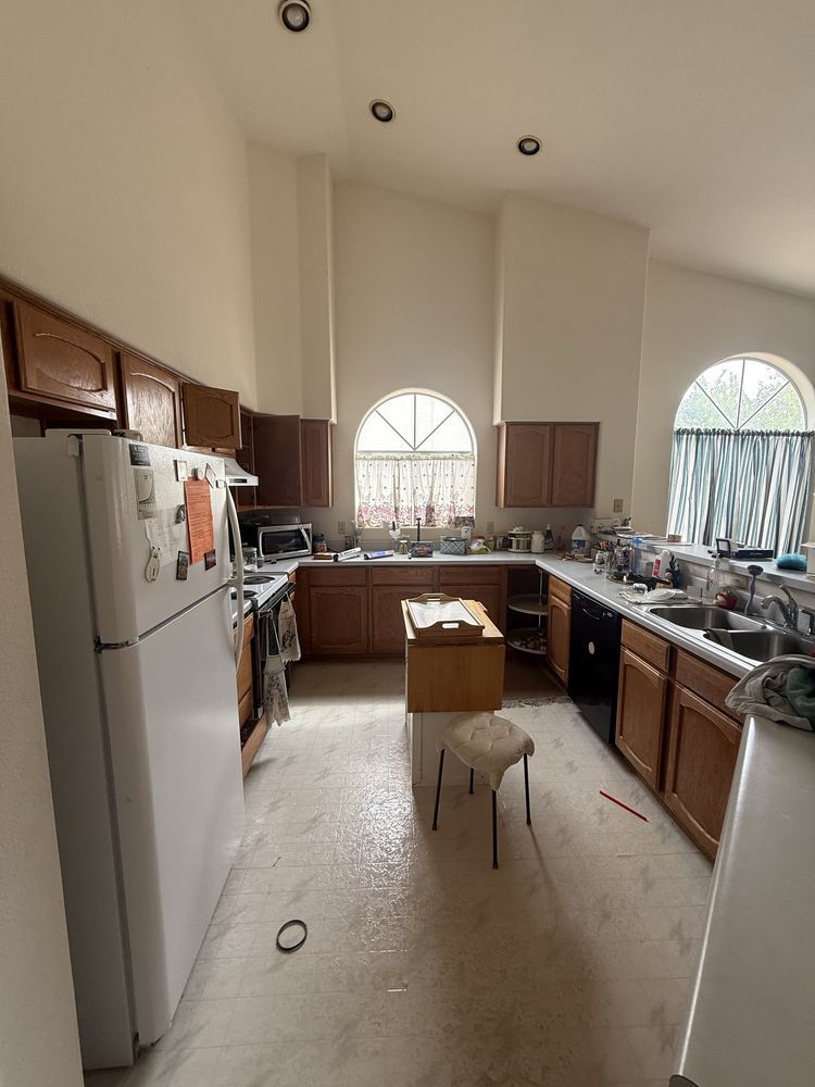 Bright kitchen with white refrigerator, wooden cabinets, arched windows, and a small stool in the center.