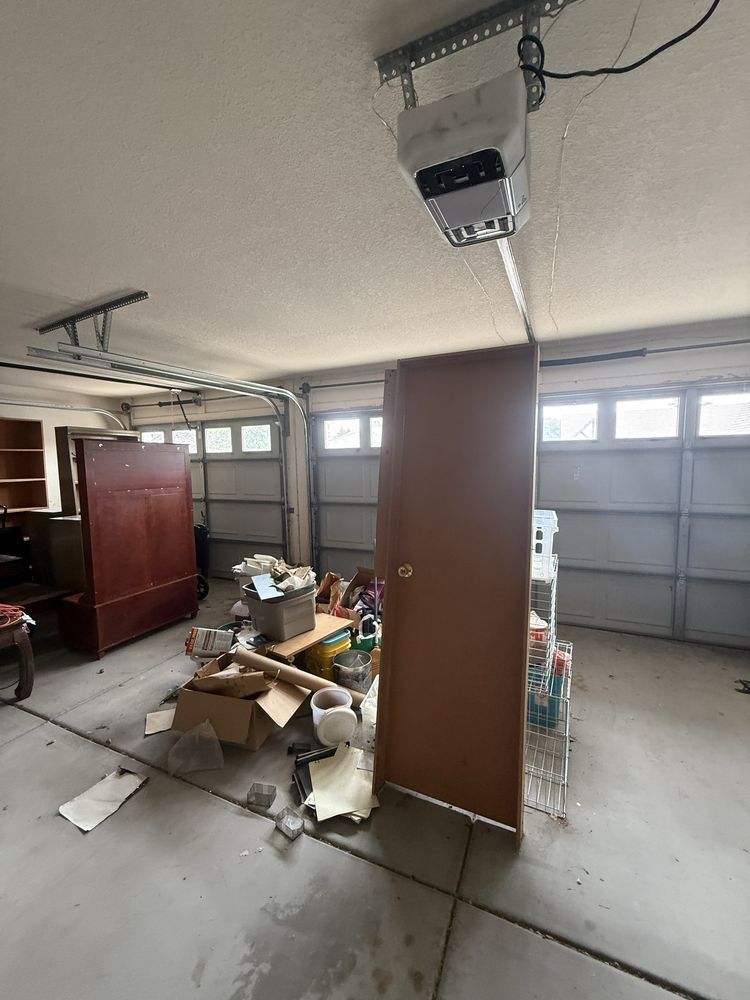 Cluttered garage with scattered debris, cabinets, and a ceiling-mounted opener under windows and a closed door.