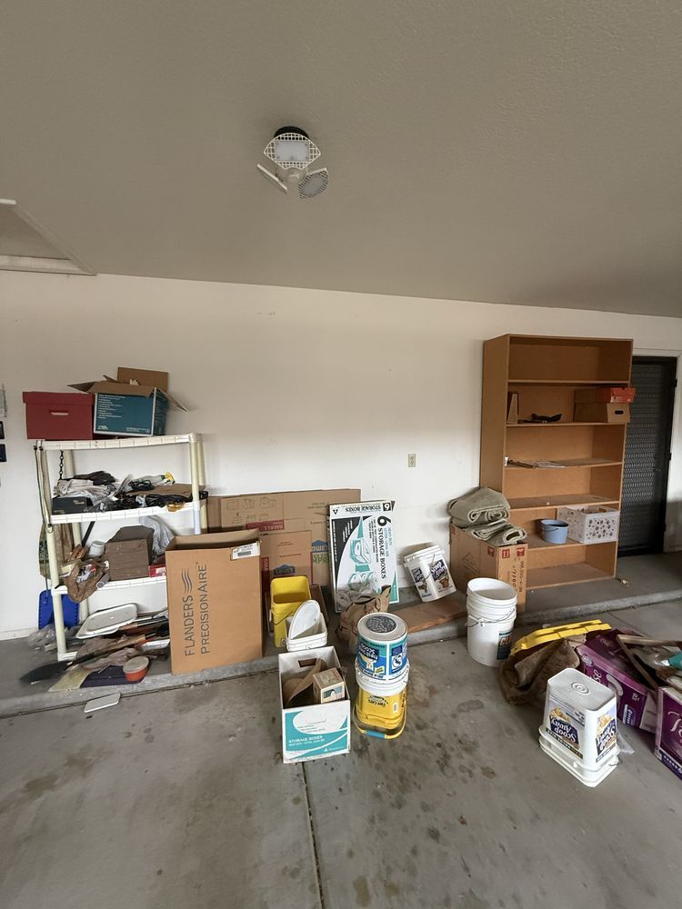 Cluttered garage with stacked boxes, tools, paint cans, and shelves along the wall