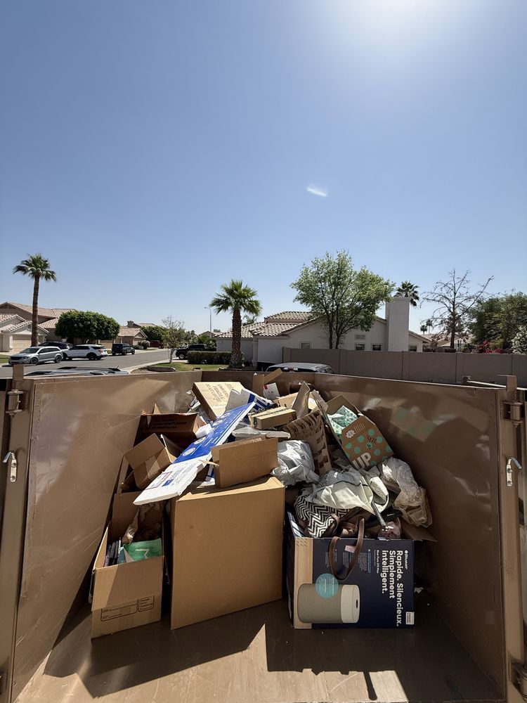 Overflowing cardboard and trash bags in a dumpster under a bright blue sky, with houses and palm trees behind.