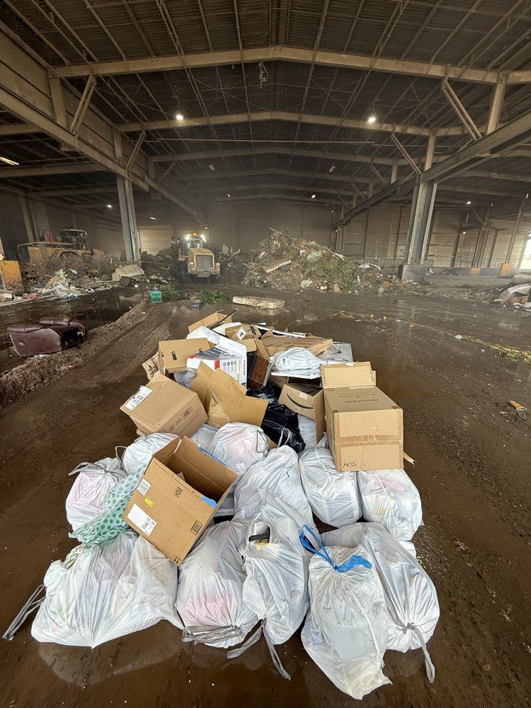 Large warehouse interior with piles of trash bags and cardboard boxes strewn across a dirty floor.
