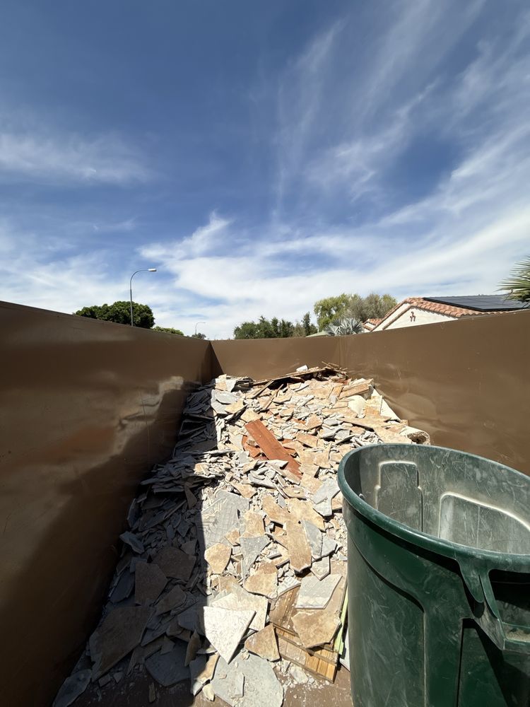 Dumpster full of broken tile and debris under a blue sky, with a green trash bin in front