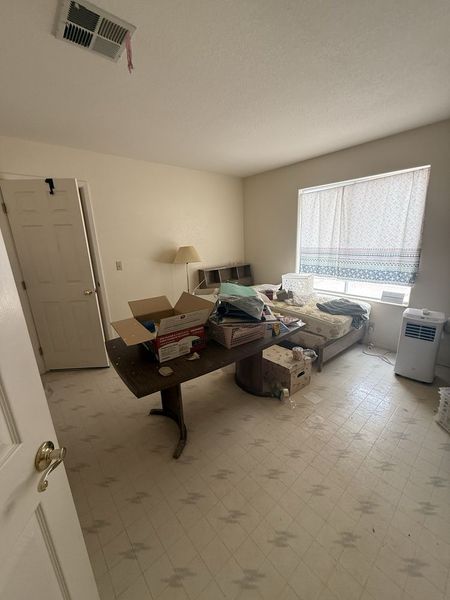 Small cluttered bedroom with bed, table, boxes, lamp, and a window with blinds.