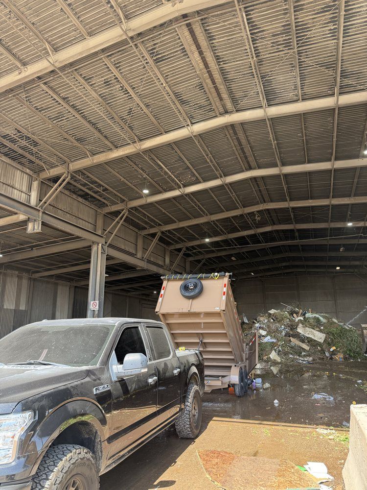 Black pickup truck beside a tan dump truck inside a warehouse or garage with a corrugated metal roof