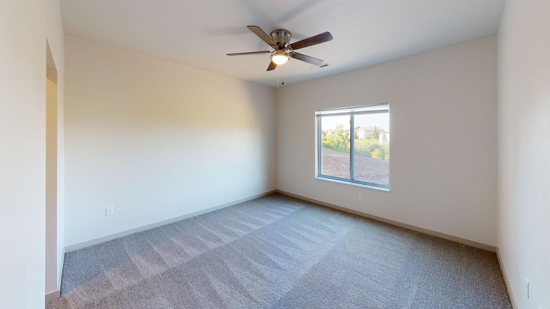 Empty beige bedroom with gray carpet, ceiling fan, and a window letting in daylight