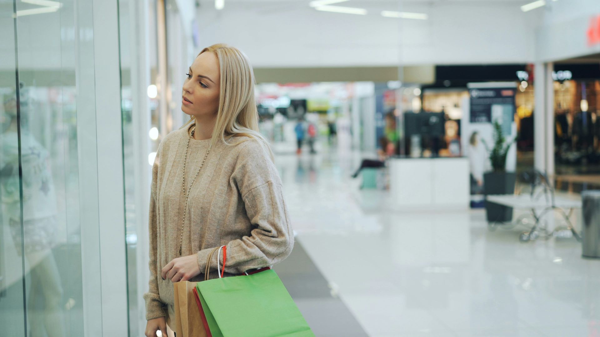 Person in beige sweater shopping in a bright mall, holding a green shopping bag and looking at a store window.