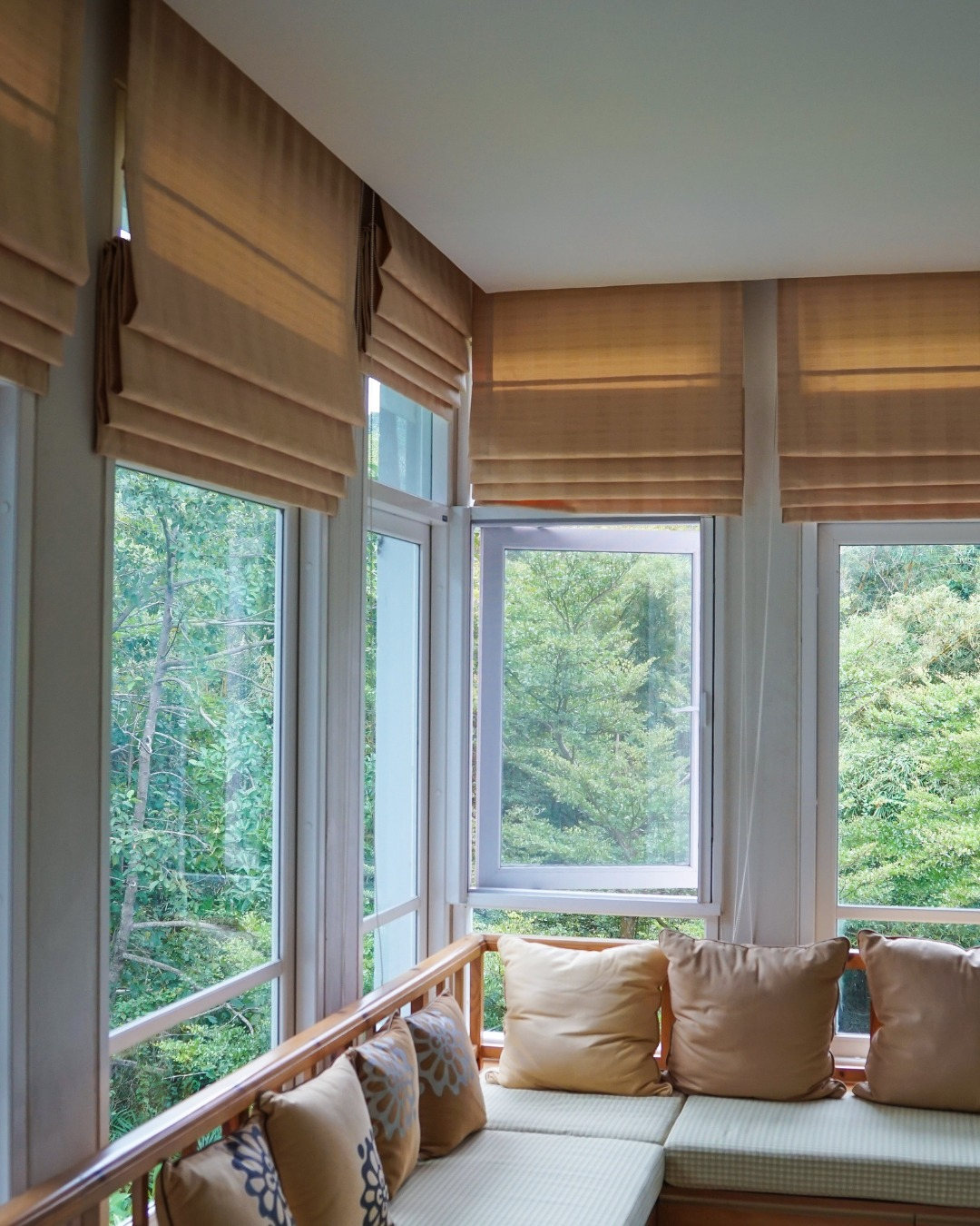 Bright sunroom corner with large windows, woven shades, and a cushioned bench overlooking green trees