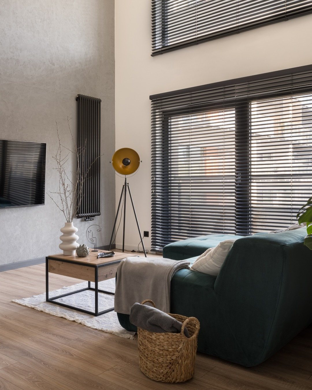 Modern living room with a dark green sofa, wooden coffee table, floor lamp, wall art, and large window blinds