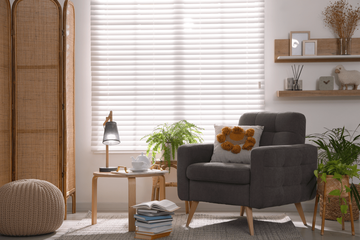 Cozy living room with gray armchair, side table, plants, wicker screen, and bright window blinds