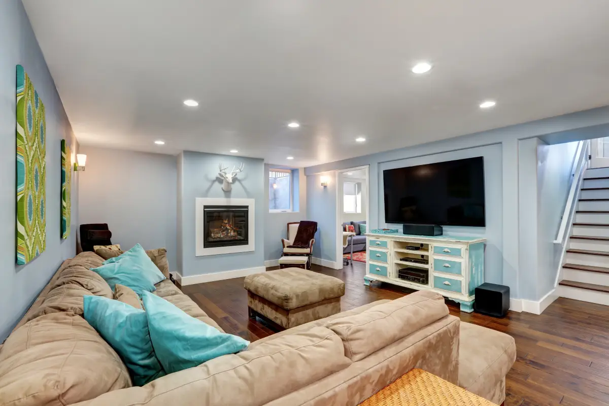 Bright living room with beige sectional, turquoise pillows, fireplace, wall TV, and staircase.