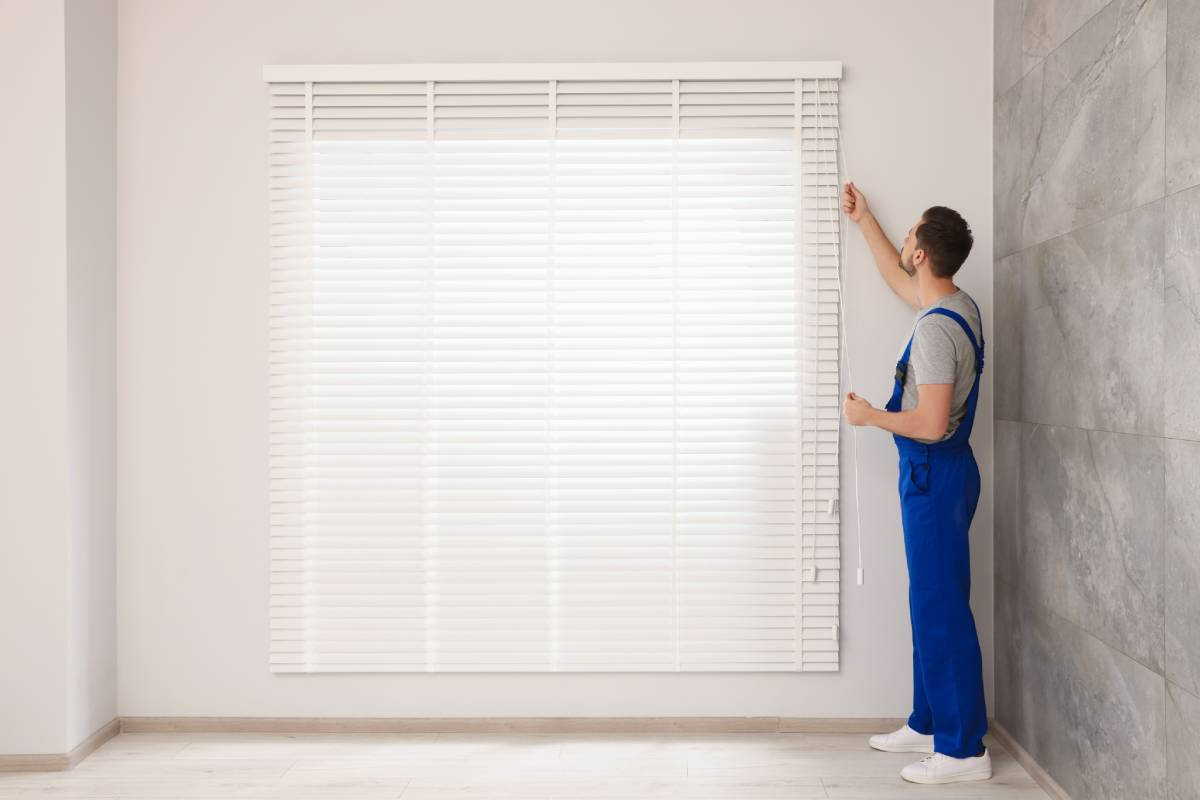 Person in blue overalls adjusting a white window blind in a bright room