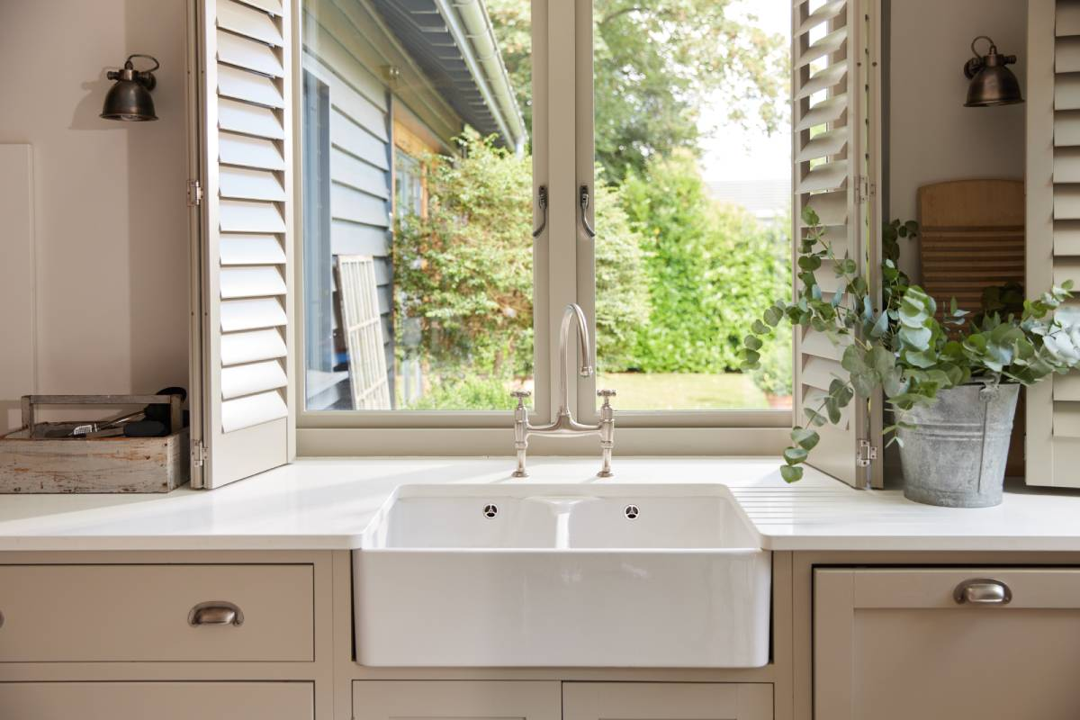 Kitchen sink beneath open window with white cabinets and potted plants on the counter