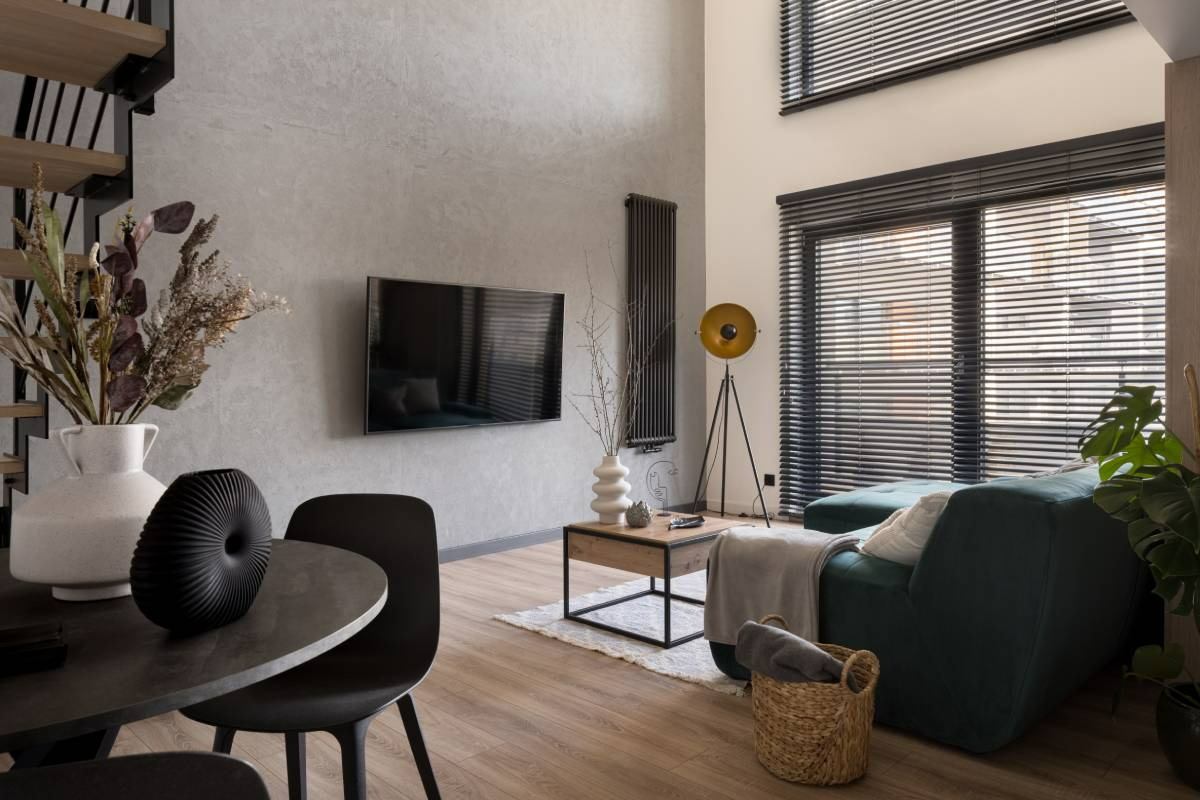 Modern living room with wall-mounted TV, large windows, dark chair, dining table, and green sofa.
