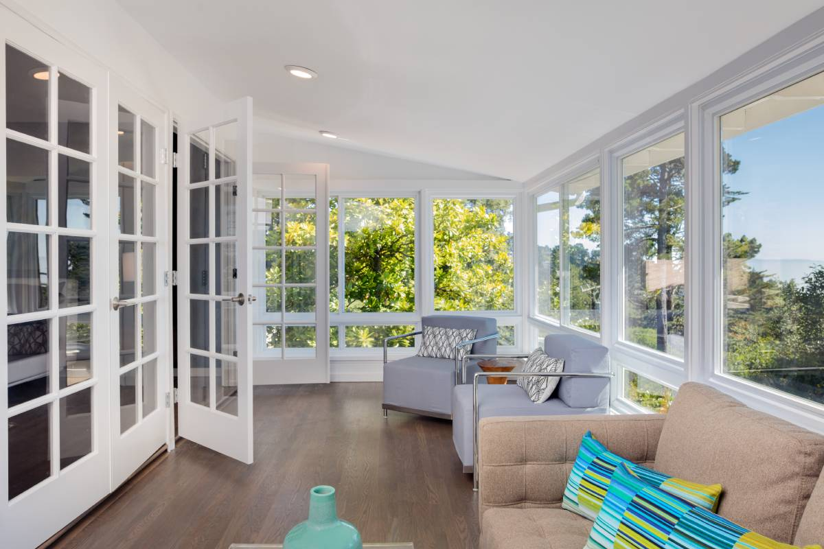 Bright sunroom with white French doors, gray sofa, tan chair, and large windows overlooking greenery
