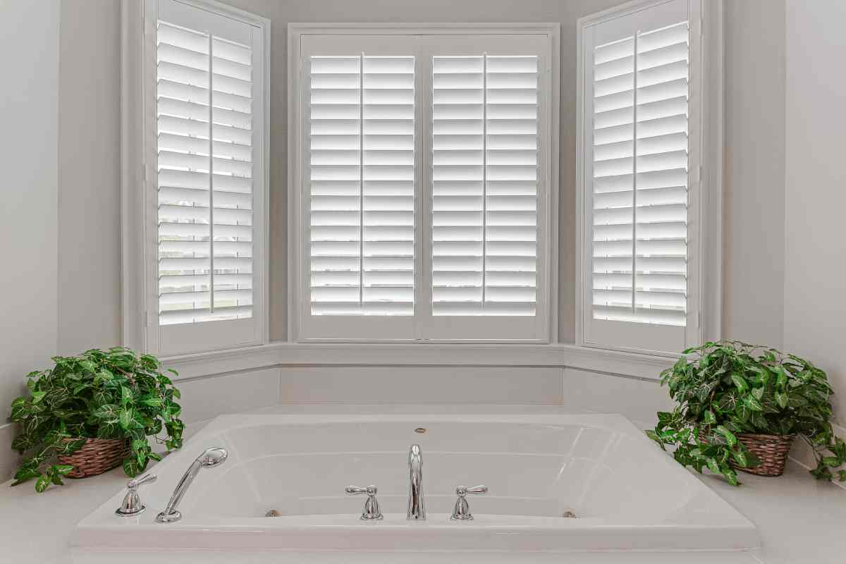 Bright bathroom with a white soaking tub beneath shuttered windows and potted plants on both sides