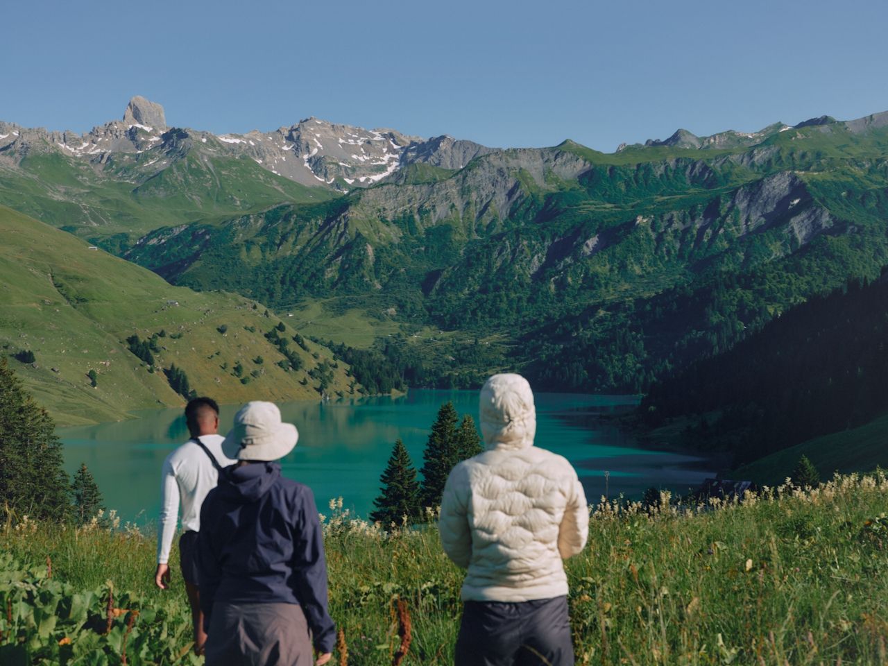 Three hikers overlook a turquoise mountain lake with green slopes and rocky peaks.