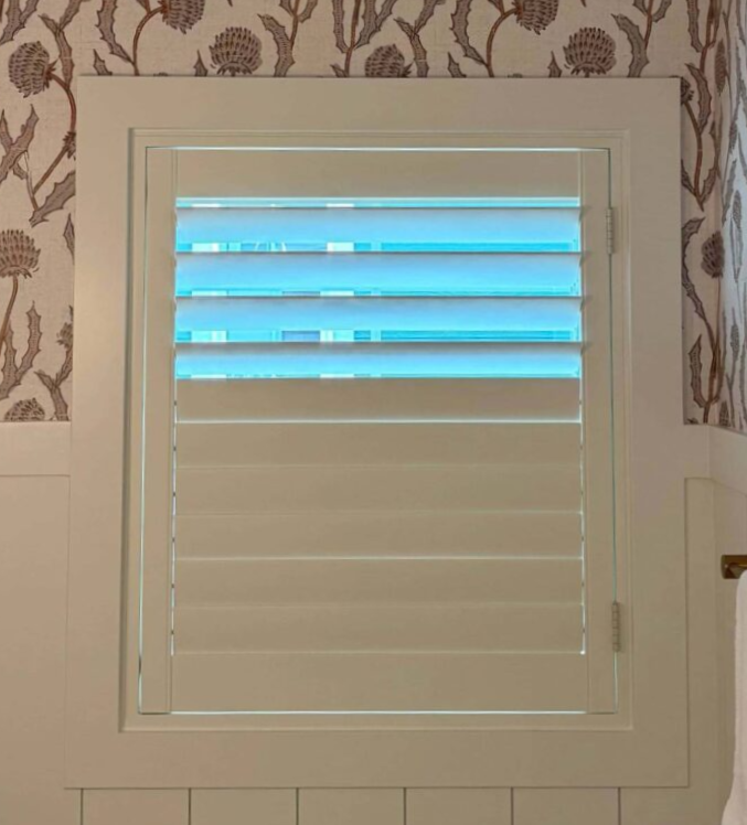 White plantation shutter with upper slats open, letting blue daylight into a patterned room.