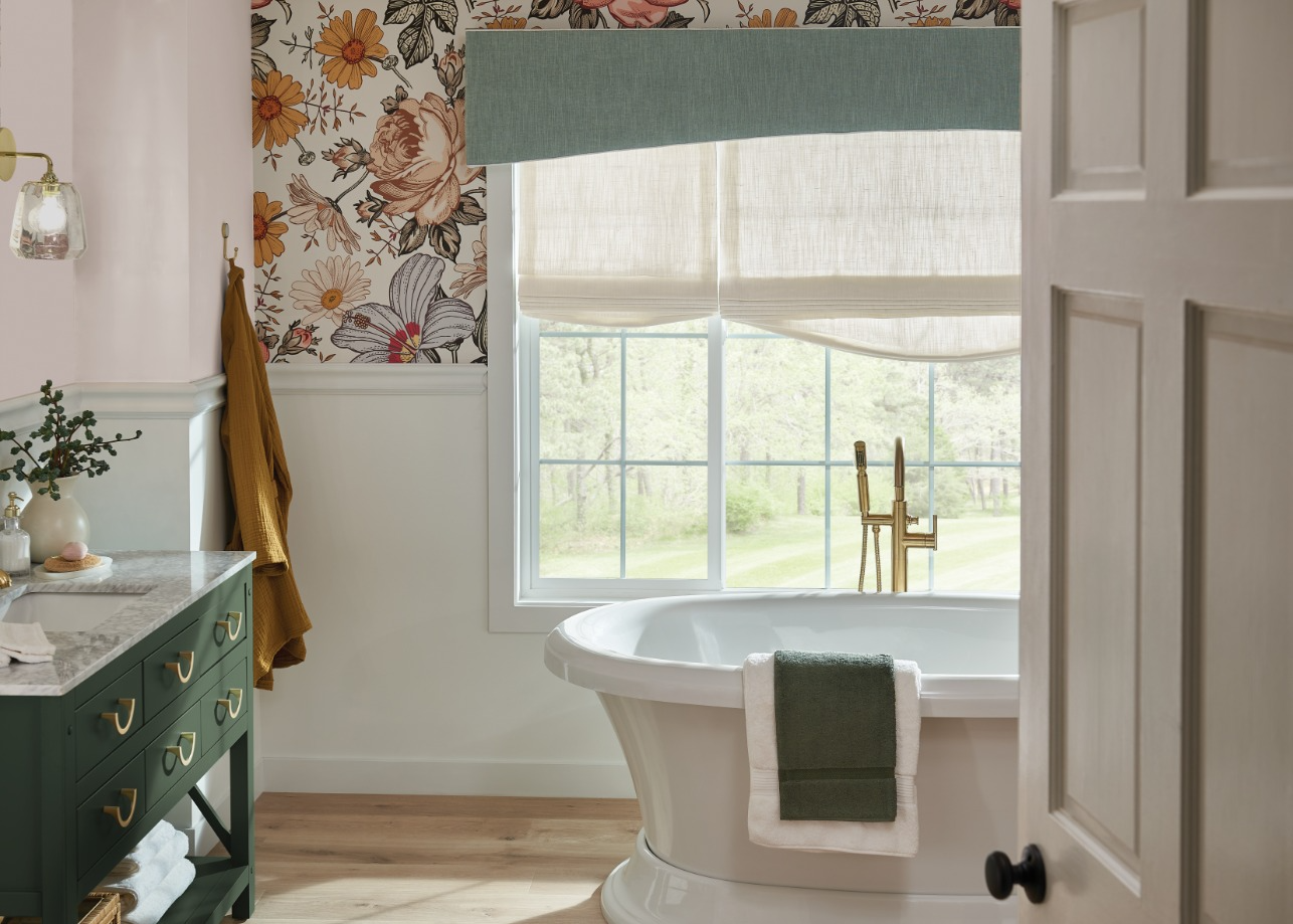 Bright bathroom with floral wallpaper, green vanity, window, and freestanding tub with towel.