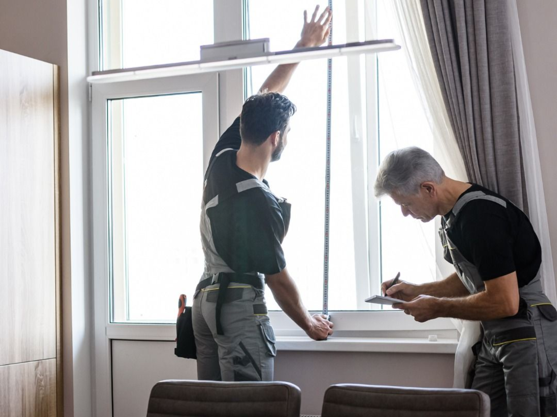 Two workers measuring a bright window opening in a room with curtains and a ladder