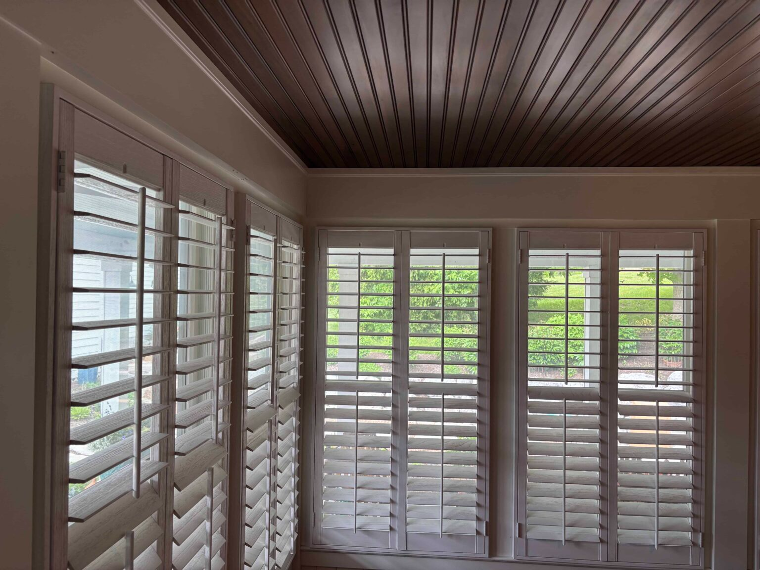 Bright room with white plantation shutters and slatted wood ceiling, looking out to green trees.