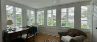 Bright living room with white shutters, a desk by the windows, and a cozy armchair near a bay window.