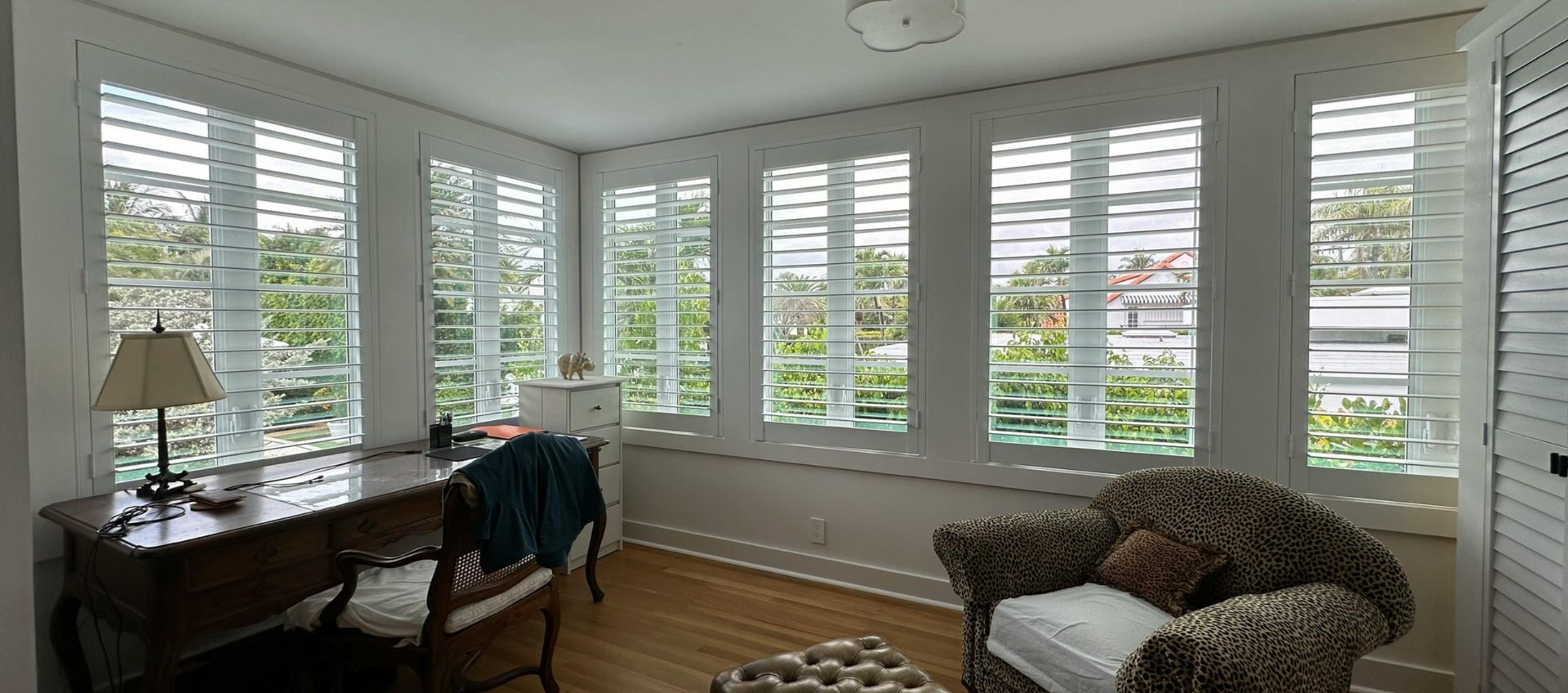 Bright living room with white shutters, a desk by the windows, and a cozy armchair near a bay window.