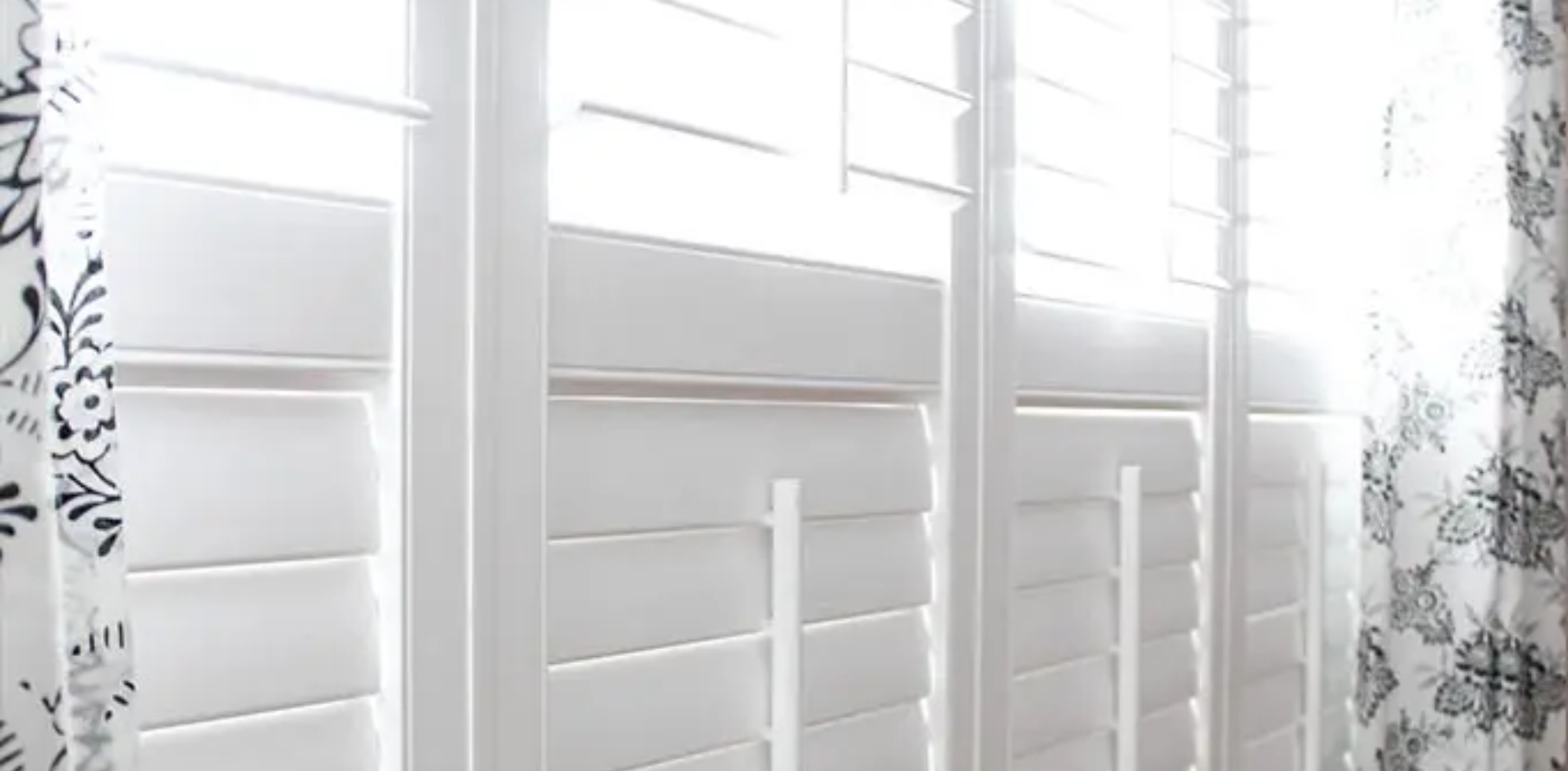 White plantation shutters filtering bright daylight beside a sheer floral curtain