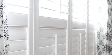 White plantation shutters with sunlight filtering through, beside a sheer floral curtain