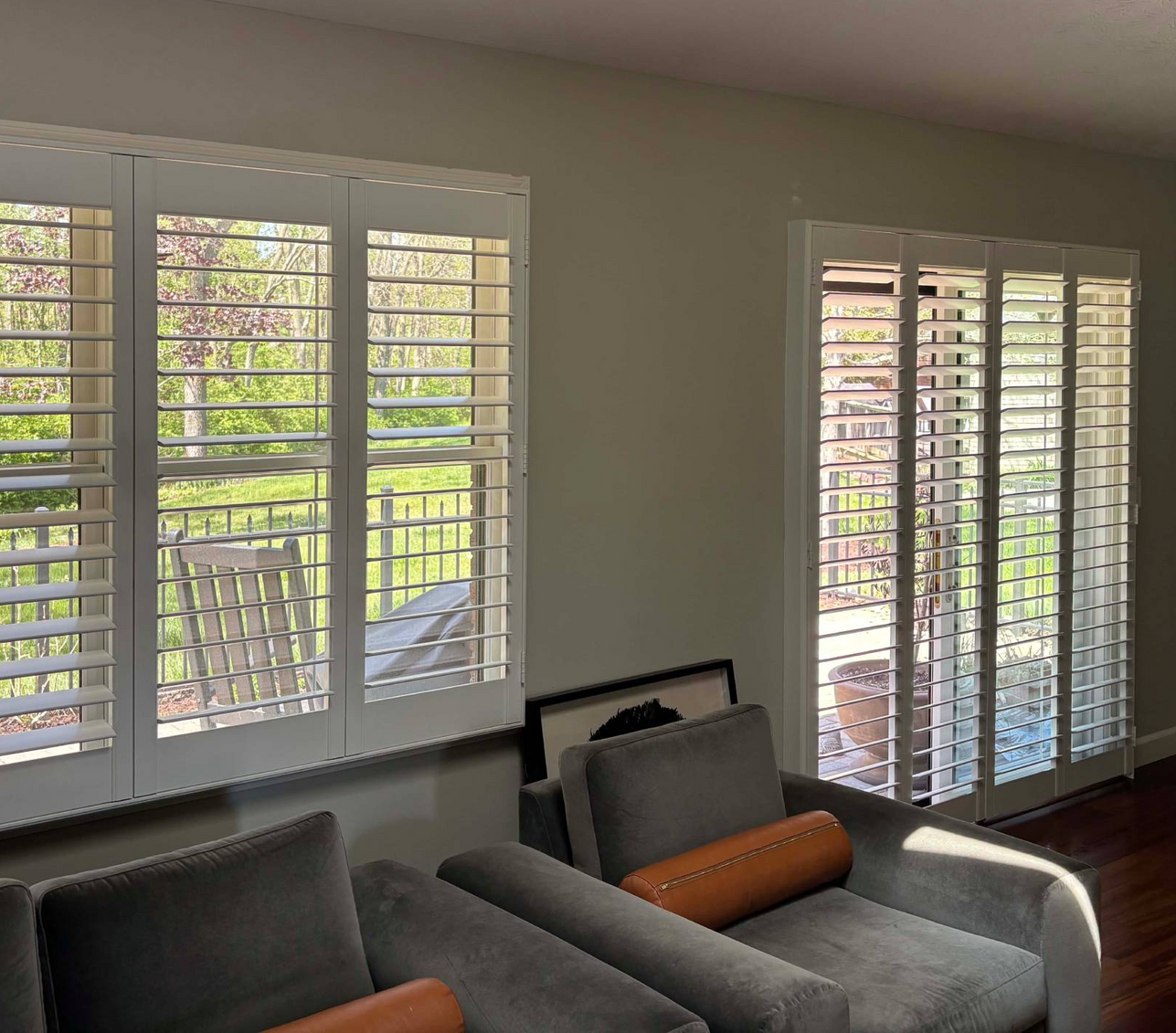 Bright living room with white shutters, sunlight, and gray chairs by large windows.