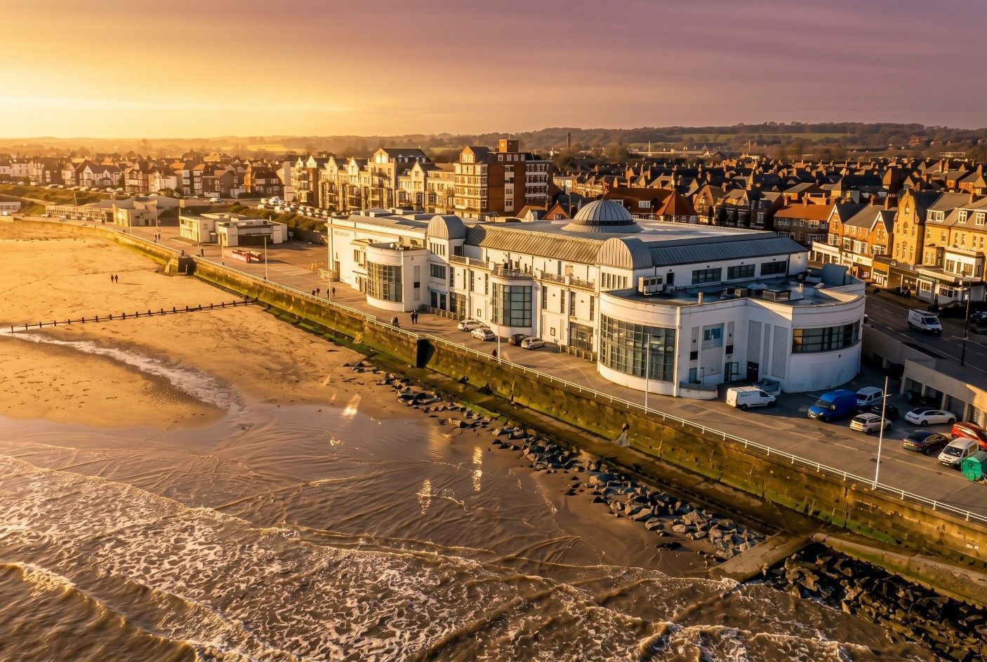 Aerial view of a seaside town with a white waterfront building, beach, and sunset-lit shoreline