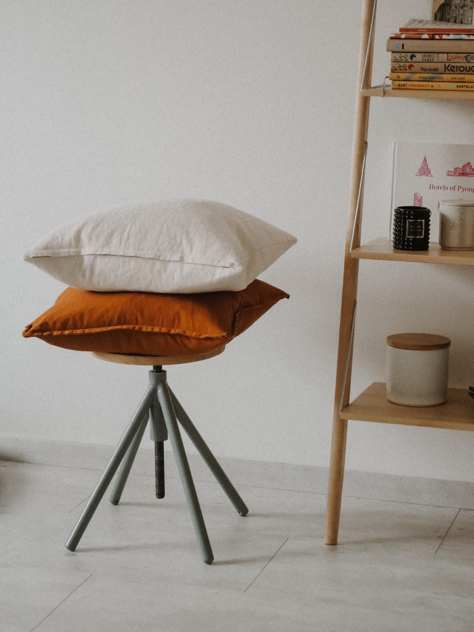 Two stacked pillows on a small tripod stool beside a wooden shelf in a minimalist room