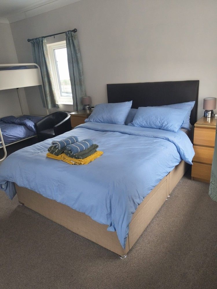 Neatly made bed with blue bedding in a gray bedroom, with a lamp, window, and folded towels.