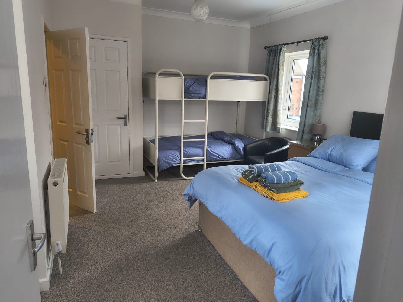 Bright bedroom with blue bedding, bunk bed, carpet, and open door near a window.