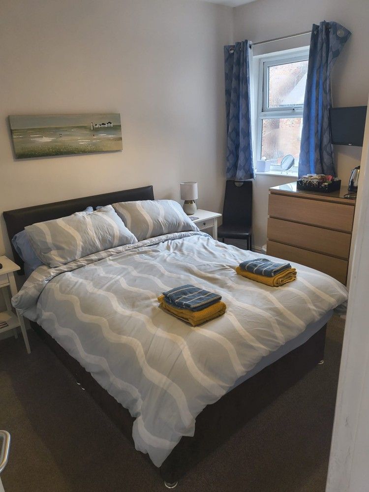 Neatly made bed in a small bedroom with blue curtains, a dresser, and folded towels on top