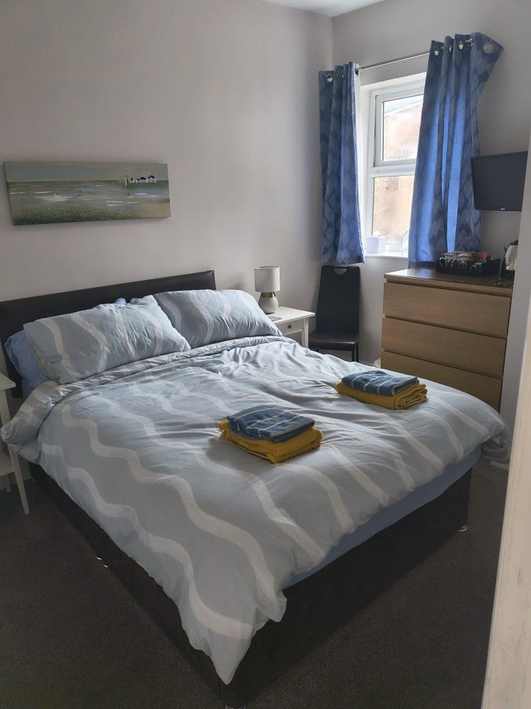 Neatly made bed in a bright bedroom with blue curtains, dresser, and wall art.