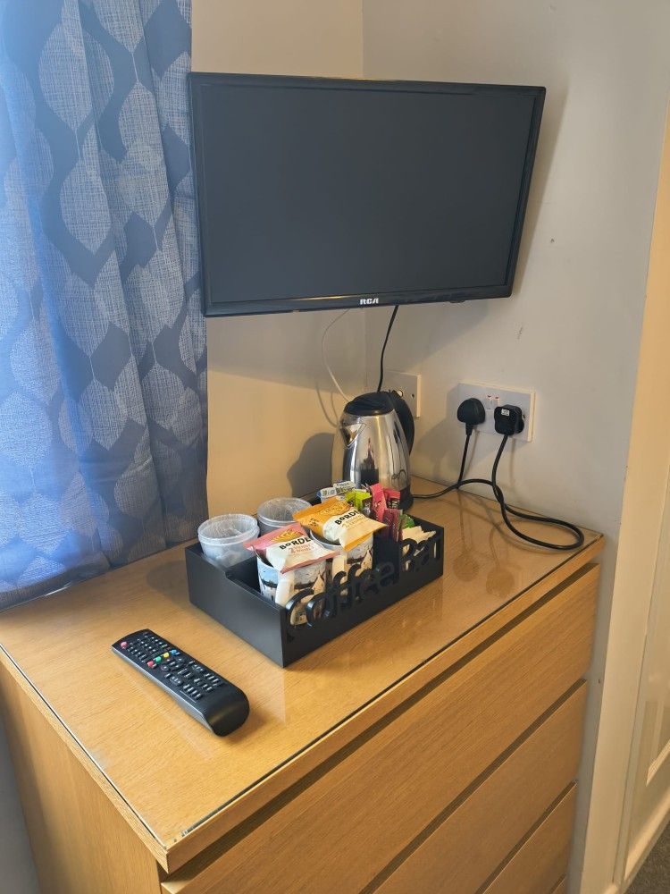 Small wall-mounted TV above a dresser with a remote, tissue box, and kettle beside a window curtain