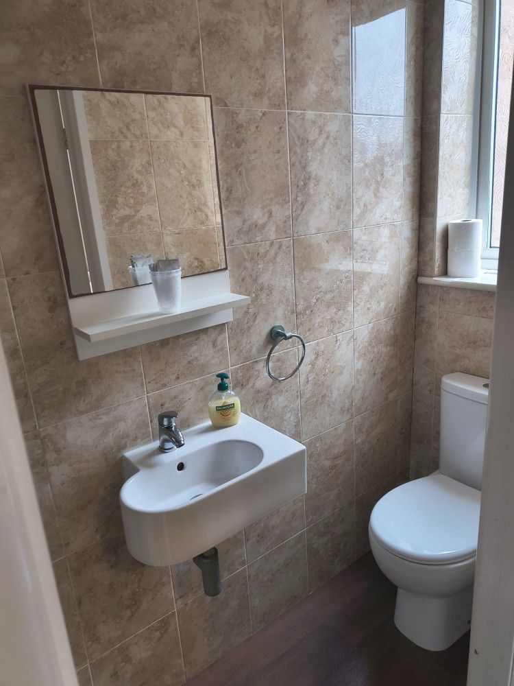 Small beige-tiled bathroom with wall-mounted sink, mirror, toilet, and window ledge with toiletries.