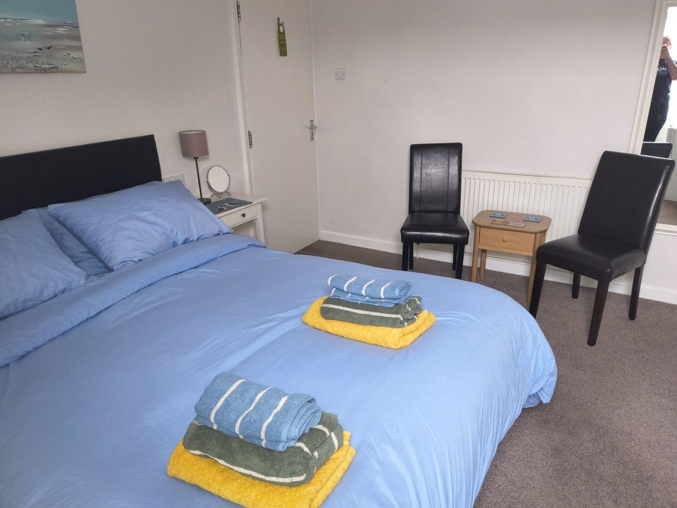 Neatly made blue bed with folded towels, two chairs, and a small table in a simple bedroom.