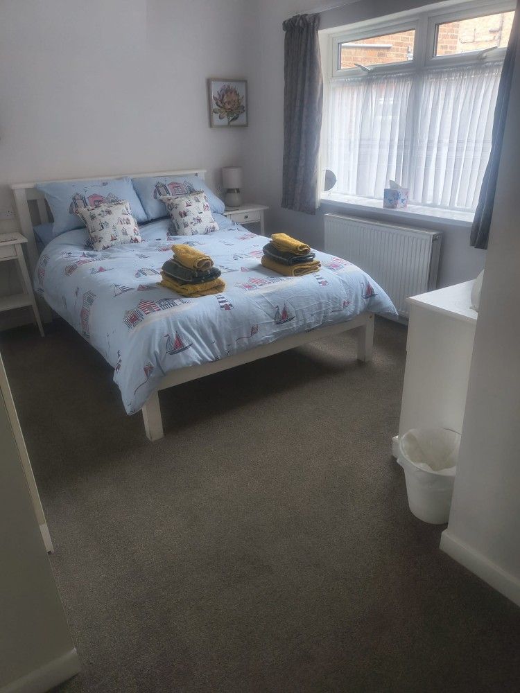 Neatly made bed in a bright bedroom with floral bedding, towels folded at the foot, and a window beside it.