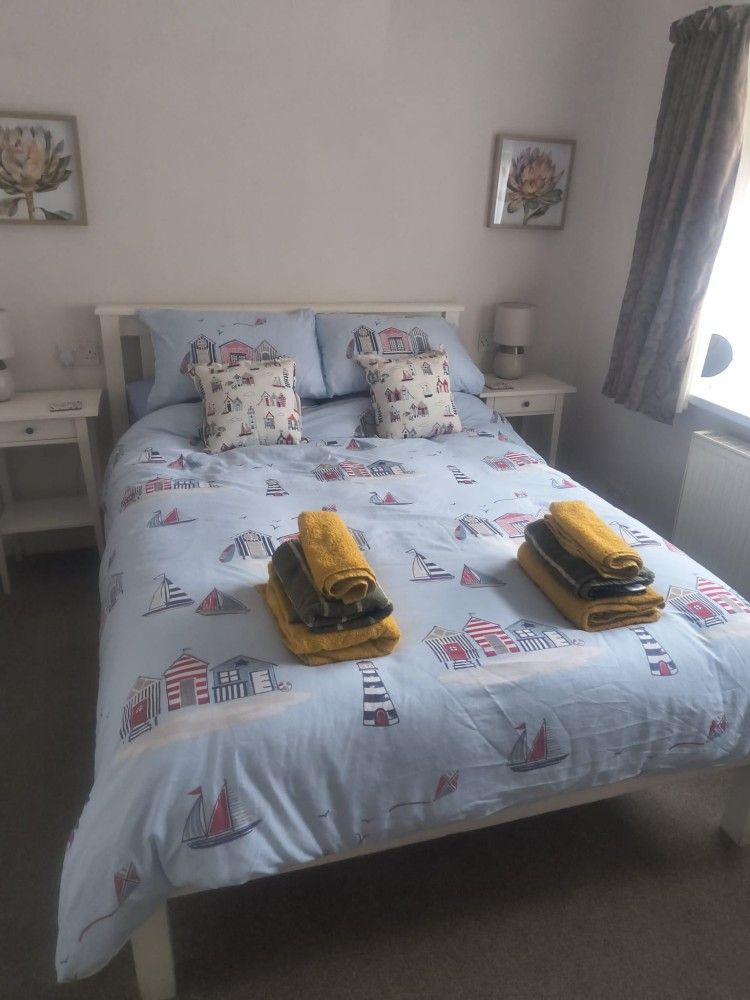 Neatly made bedroom with a blue patterned bedspread, folded yellow towels, bedside tables, and framed wall art.