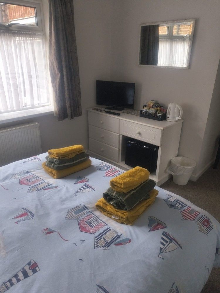 Bedroom with a neatly made bed, folded yellow towels, a white dresser, mirror, TV, and window with curtains.