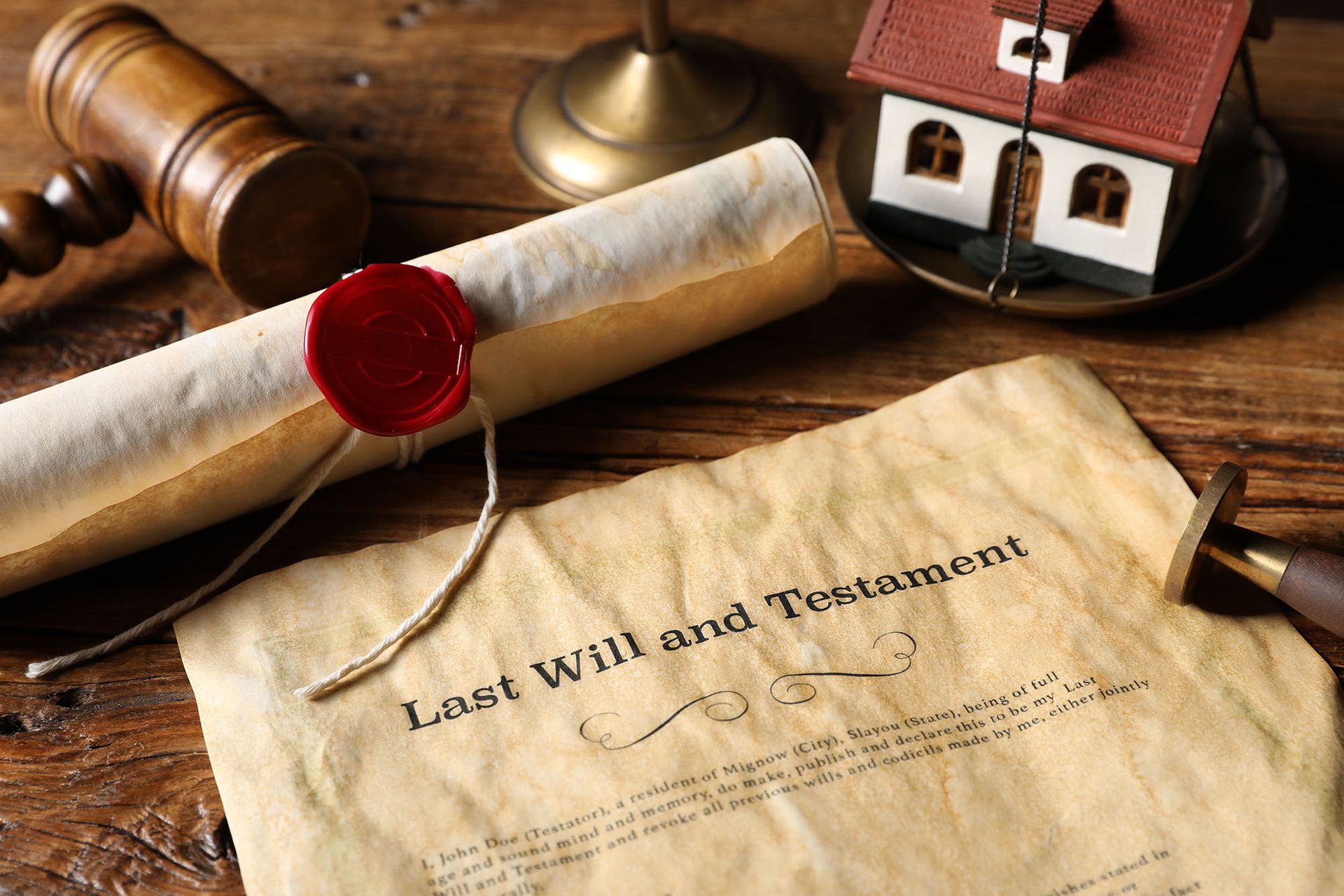 A wooden gavel, a model house, and a rolled-up document with a red wax seal rest on a table next to a last will document.